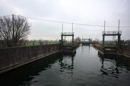 Sluices of a dam, seen from behind a diversionary channel above an embankment, with the sky cast on...
