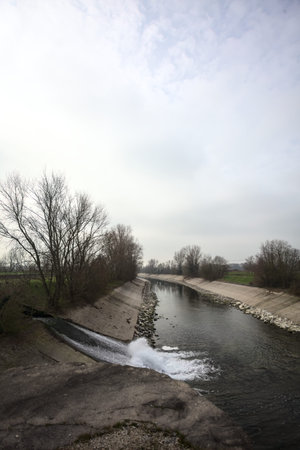 Diversionary channel with rows of bare trees over its banks on a cloudy day in the Italian...