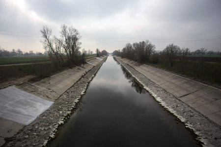 Diversionary channel with bare trees bordering the banks, reflected in the water together with the sky, on a cloudy dayの写真素材