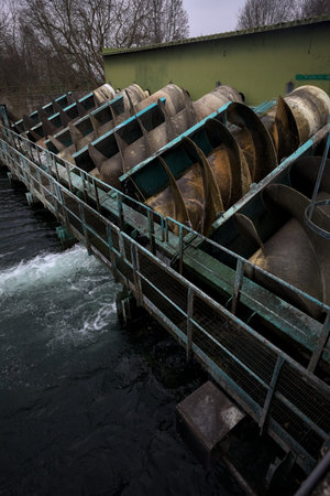 Moving water screw among still others, with water splashing next to it, framed by a footbridge, in a...
