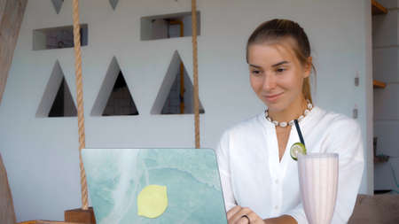 Photo of a young girl sitting at a wooden table and working behind a laptop in a green gardenの写真素材