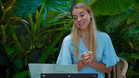 Photo of a young girl sitting at a wooden table and working behind a laptop in a green gardenの写真素材