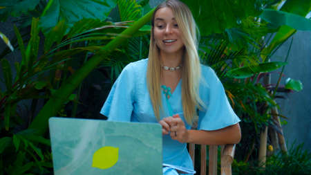 Photo of a young girl sitting at a wooden table and working behind a laptop in a green gardenの写真素材
