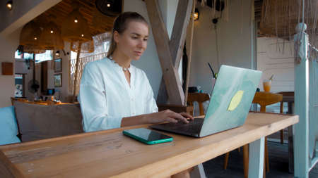 Photo of a young girl sitting at a wooden table and working behind a laptop in a green gardenの写真素材