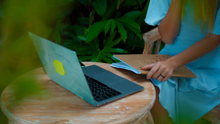 Photo of a young girl sitting at a wooden table and working behind a laptop in a green gardenの写真素材