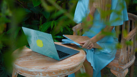 Photo of a young girl sitting at a wooden table and working behind a laptop in a green gardenの写真素材