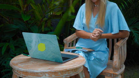 Photo of a young girl sitting at a wooden table and working behind a laptop in a green gardenの写真素材