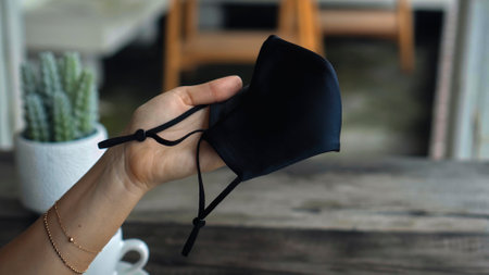 A young European girl sits at a table in a public place and holding a black protective mask with her handsの写真素材