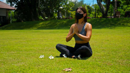 Young European brunette girl practices alone yoga in nature sitting on green grass in lotus pose wearing a black protective mask on her faceの写真素材