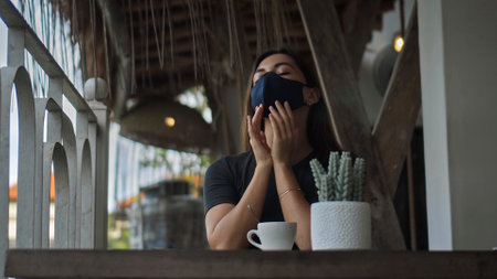 A young European brunette girl sits at a table in a public place and touches a soft black protective mask with her hands on her faceの写真素材