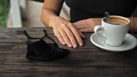 A young European girl sits at a table in a public place with a cup of coffee and nearby to her on a wooden table is a black protective maskの写真素材