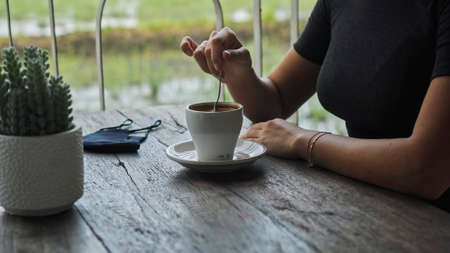 A young European girl sits at a table in a public place with a cup of coffee and nearby to her on a wooden table is a black protective maskの写真素材