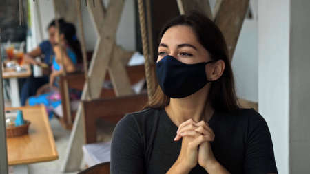 A young European brunette girl sits at a table in a public place on a black protective mask on her...