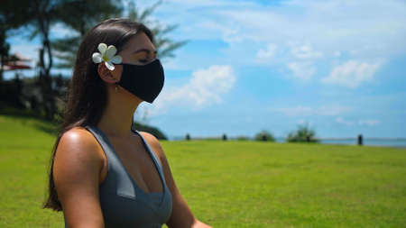 A young European brunette girl practices alone yoga in nature sitting on green grass in a lotus pose wearing a black protective mask on her face and a white flower behind her earの写真素材