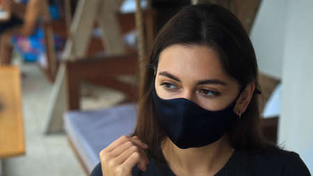 A young sad European brunette girl sits at a table in a public place on a black protective mask on her faceの写真素材