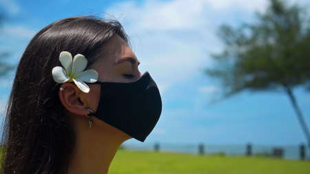 A young European brunette girl practices alone yoga in nature sitting on green grass in a lotus pose wearing a black protective mask on her face and a white flower behind her earの写真素材