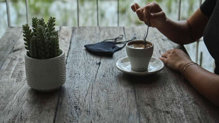 A young European girl sits at a table in a public place with a cup of coffee and nearby to her on a wooden table is a black protective maskの写真素材