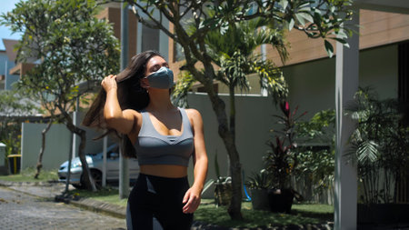 A young European brunette sports girl walks along an empty city street with green trees on the background alone in a gray protective mask on her faceの写真素材