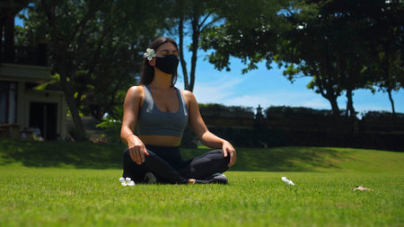 Young European brunette girl practices alone yoga in nature sitting on green grass in lotus pose wearing a black protective mask on her faceの写真素材
