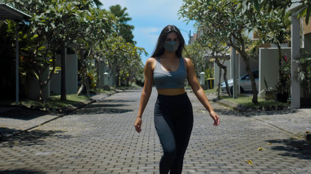 A young European brunette sports girl walks along an empty city street with green trees on the background alone in a gray protective mask on her faceの写真素材