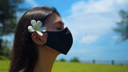 A young European brunette girl practices alone yoga in nature sitting on green grass in a lotus pose wearing a black protective mask on her face and a white flower behind her earの写真素材