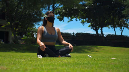 Young European brunette girl practices alone yoga in nature sitting on green grass in lotus pose wearing a black protective mask on her faceの写真素材