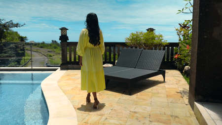 Young long-haired girl walking in the backyard with a green garden, pool with blue clean water and blue sky in a beautiful long yellow dressの写真素材