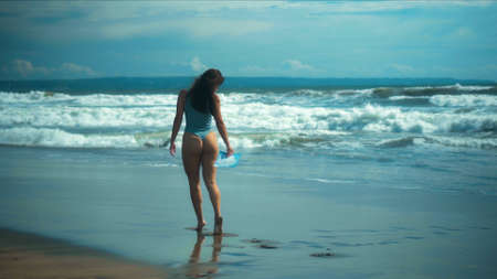 A young sexy European girl in a bikini walks on black sand by the sea with blue wavesの写真素材
