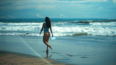 A young sexy European girl in a bikini walks on black sand by the sea with blue wavesの写真素材