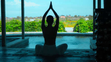 A young European brunette girl sits on a towel and yoga in a lounge room overlooking green fieldsの写真素材