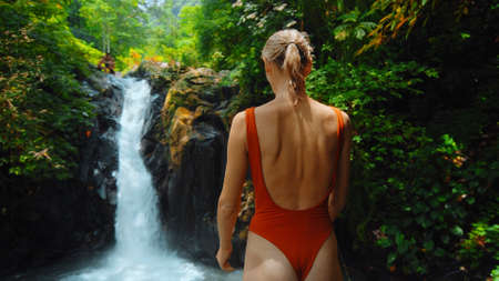 waterfall with rocks among tropical jungle with green plants and trees and water falling down into river with woman staying near the waterfallの写真素材