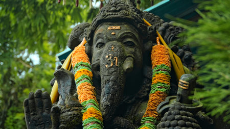 An old stone statue of Ganesha covered with moss with a wreath of yellow flowers on his neck standing on the street with green plants on the backgroundの写真素材