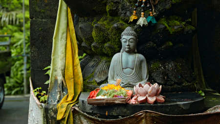 An old stone Budist statue sitting in a lotus pose covered with moss with a wreath of yellow flowers on its neck standing on the street with green plants on the backgroundの写真素材