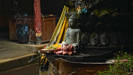 An old stone Budist statue sitting in a lotus pose covered with moss with a wreath of yellow flowers on its neck standing on the street with green plants on the backgroundの写真素材