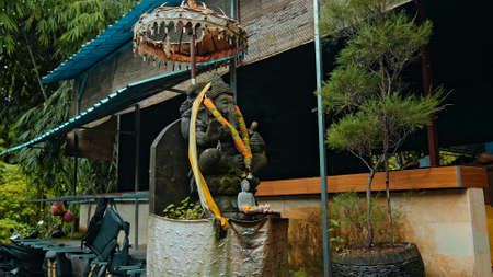 An old stone statue of Ganesha covered with moss with a wreath of yellow flowers on his neck standing on the street with green plants on the backgroundの写真素材