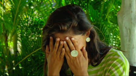 A young European brunette girl sits in the backyard smiling and touches herself with her hands for her face with green plants on the backgroundの写真素材