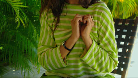 A young girl in a green dress with made a heart with her hand sitting on a bench in the backyard with green plants on the backgroundの写真素材