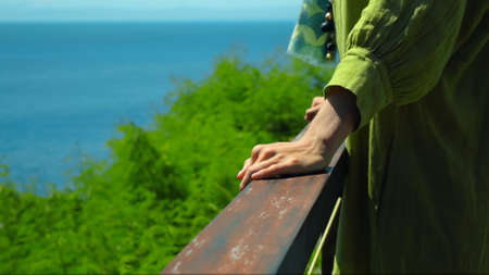 A young European girl stands in the summer on a balcony holding her hand to a wooden railing in a green dressの写真素材