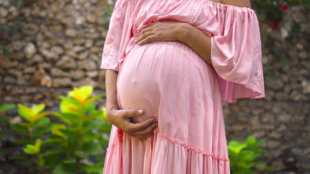 A young European pregnant girl in a pink dress gently touches her big stomach with her hands, stands on the back green gardenの写真素材