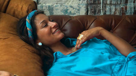A beautiful young European brunette girl in a blue dress lies on the couch and touches her hands with a golden ringの写真素材