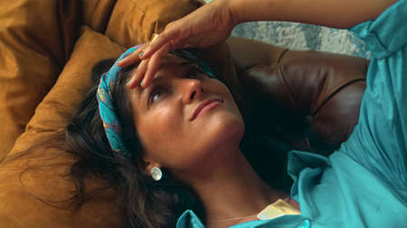 A beautiful young European brunette girl in a blue dress lies on the couch and touches her hands with a golden ringの写真素材