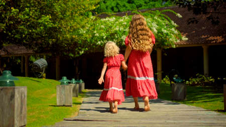 Mother and daughter walking in the backyard with a green garden and grass in a beautiful long red dresses, turning her backの写真素材