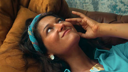 A beautiful young European brunette girl in a blue dress lies on the couch and touches her hands with a golden ringの写真素材