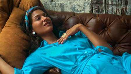 A beautiful young European brunette girl in a blue dress lies on the couch and touches her hands with a golden ringの写真素材