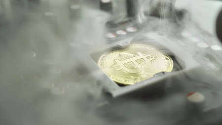 A gold Bitcoin coin with glare and reflection installed instead of a central processor in the motherboard with electronic components with light and smoke in the darkness. Bitcoin miningの写真素材