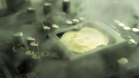 A gold Bitcoin coin with glare and reflection installed instead of a central processor in the motherboard with electronic components with light and smoke in the darkness. Bitcoin miningの写真素材