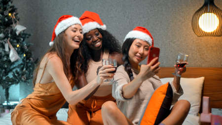 Three young girls European, Asian and African in red New Year's clothes and hats sit on a bed at a Christmas tree holding glasses of wine made photo on a phone, smiling and celebrating Christmasの写真素材