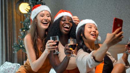 Three young girls European, Asian and African in red New Year's clothes and hats sit on a bed at a Christmas tree holding glasses of wine, smiling and celebrating Christmasの写真素材