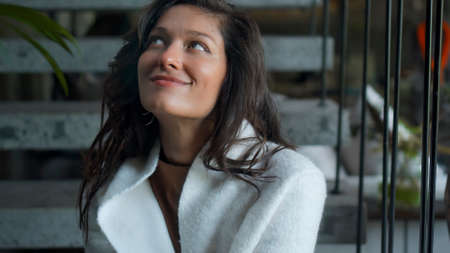 Young and beautiful European girl long-haired brunette sits at home on the stairs in autumn clothes and a white sweater and smiling touches herself with her handsの写真素材