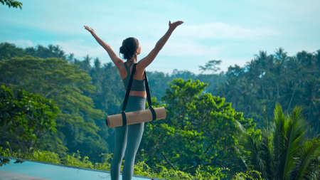 A young attractive brunette sports girl goes with a yoga rug in nature with a green rainforest on a lit background by the pool.の写真素材